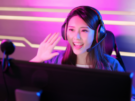 Junge Streamerin beim Gaming-Streaming sitzt vor dem Monitor, schaut in die Kamera und winkt ihren Zuschauern zu