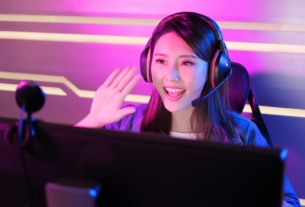 Junge Streamerin beim Gaming-Streaming sitzt vor dem Monitor, schaut in die Kamera und winkt ihren Zuschauern zu