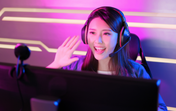 Junge Streamerin beim Gaming-Streaming sitzt vor dem Monitor, schaut in die Kamera und winkt ihren Zuschauern zu