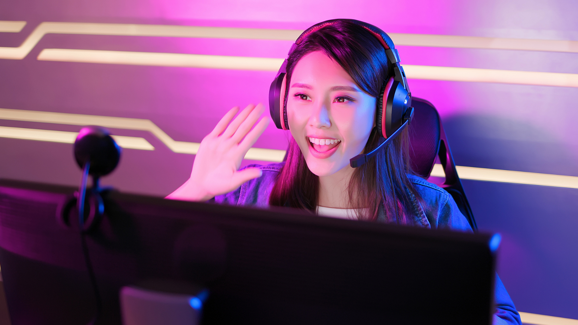 Junge Streamerin beim Gaming-Streaming sitzt vor dem Monitor, schaut in die Kamera und winkt ihren Zuschauern zu